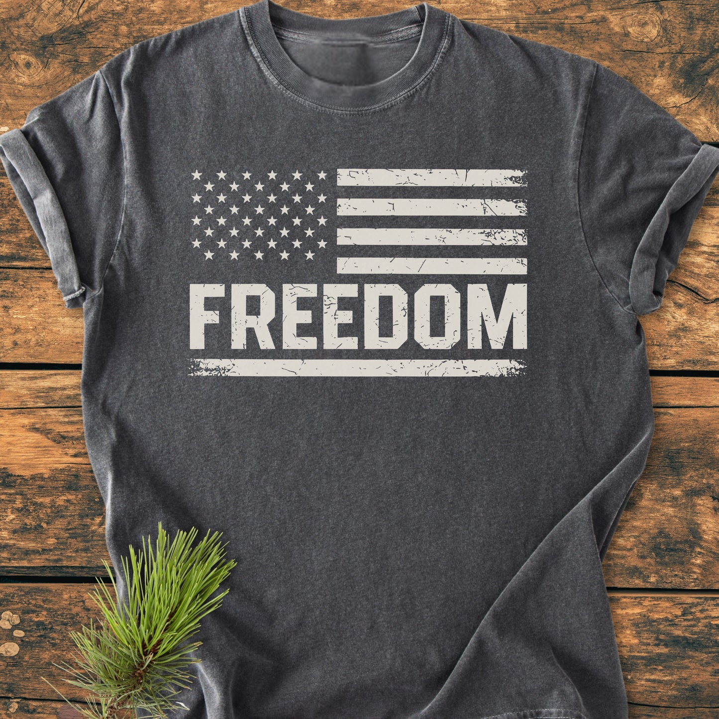 Freedom Flag - Vintage Wash Tee: Charcoal-gray T-shirt featuring a distressed white U.S. flag and FREEDOM text, laid on rustic wood with a pine sprig beside it, highlighting its relaxed, durable design.