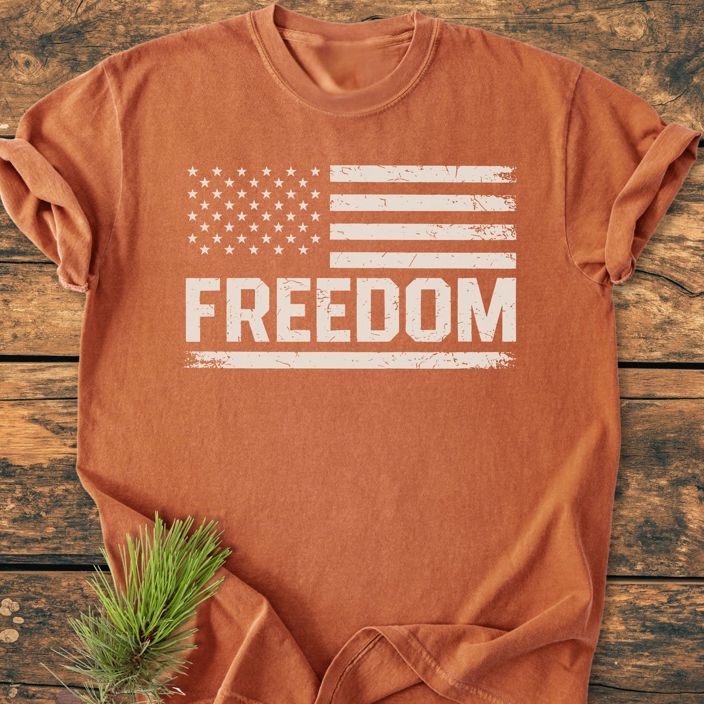Freedom Flag - Vintage Wash Tee displayed on a wooden surface, featuring a distressed U.S. flag graphic and FREEDOM text. Made from soft, garment-dyed cotton for comfort and durability.