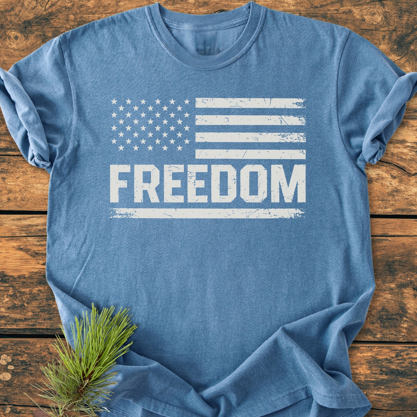 Freedom Flag - Vintage Wash Tee displayed flat with a distressed U.S. flag graphic and FREEDOM text. Made from soft, durable garment-dyed cotton, perfect for everyday wear. Pine branch accent nearby.