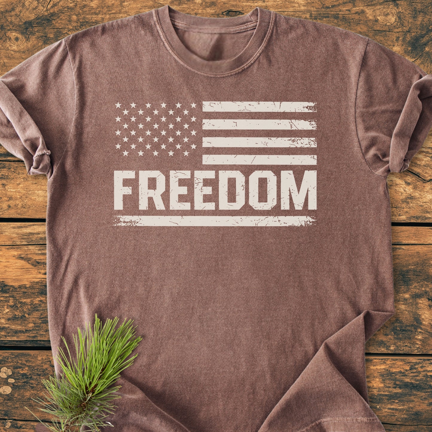 Freedom Flag - Vintage Wash Tee featuring a distressed American flag and FREEDOM text, laid flat on a rustic wooden surface with a pine branch beside it.