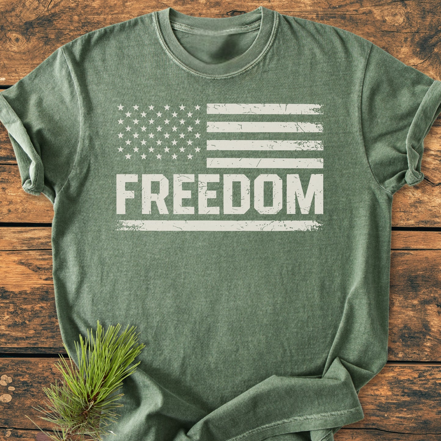 Freedom Flag - Vintage Wash Tee displayed on rustic wood, featuring a distressed U.S. flag graphic with FREEDOM text. Olive-green, soft-washed cotton with pine sprig accentuating its natural aesthetic.