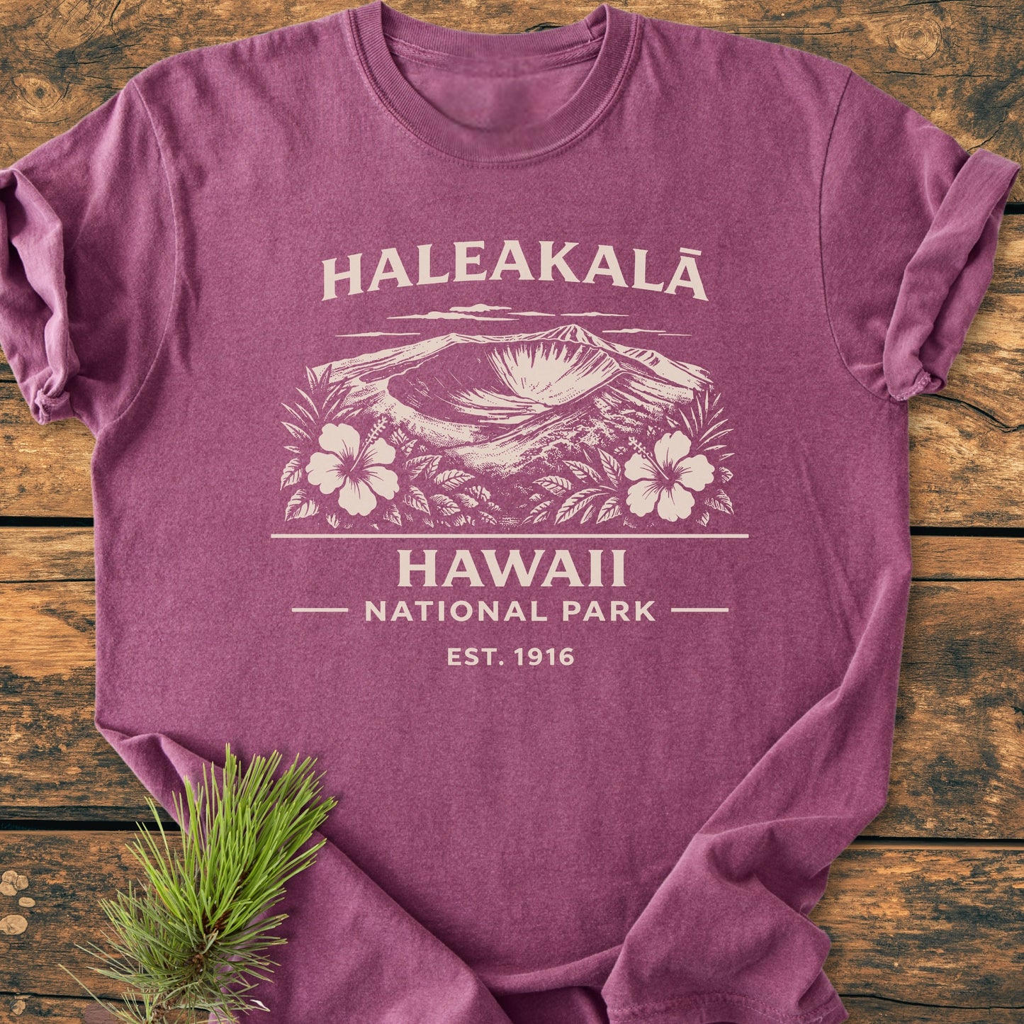 Haleakalā Hawaii National Park - Vintage Wash Tee featuring a volcanic crater graphic with tropical flowers, laid flat on wood. Includes 'HALEAKALĀ' and 'HAWAII NATIONAL PARK – EST. 1916' text, pine sprig beside.