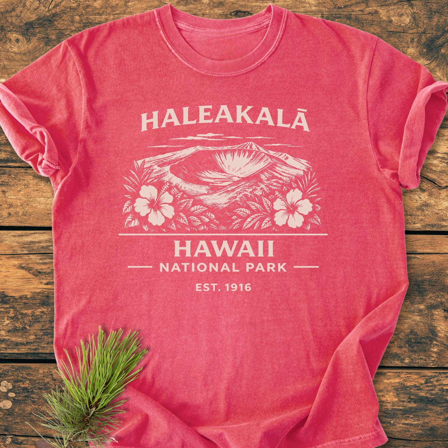Haleakalā Hawaii National Park - Vintage Wash Tee featuring a salmon-tone, white illustrated crater with hibiscus, and text. Laid on wood, pine sprig beside. Cozy, relaxed fit, 100% ring-spun cotton.
