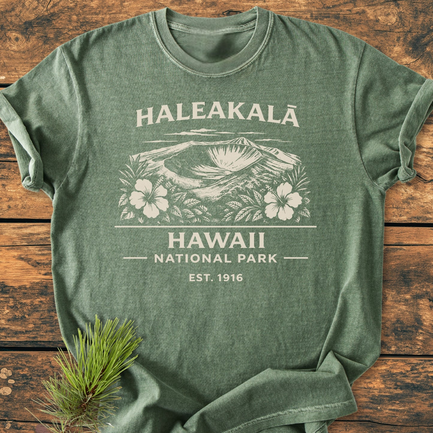 Haleakalā Hawaii National Park - Vintage Wash Tee in forest-green, featuring a white graphic of Haleakalā crater with hibiscus flowers. Displayed on a wooden surface with an evergreen sprig beside it.