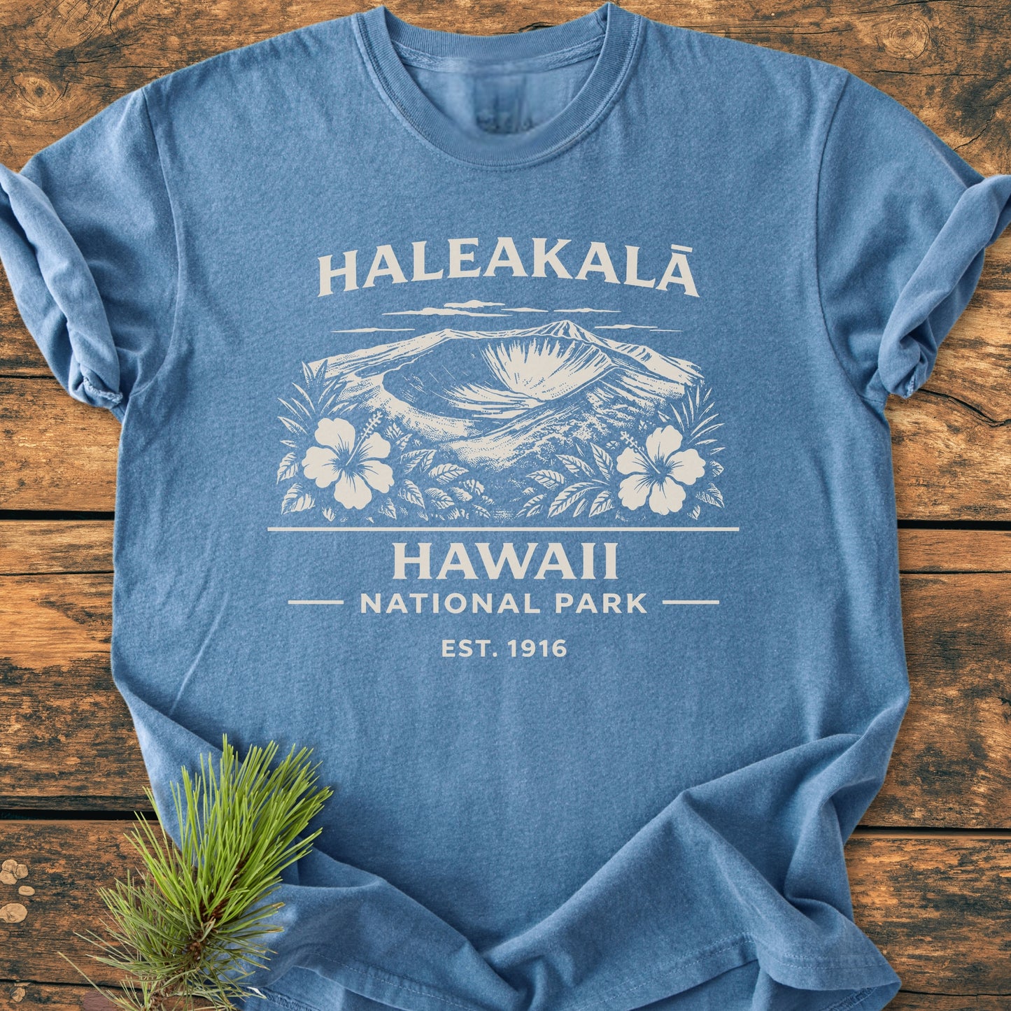 The Haleakalā Hawaii National Park - Vintage Wash Tee features a white vintage-style graphic of Haleakalā crater with hibiscus, emphasizing its relaxed fit and soft, garment-dyed fabric on a wooden backdrop.