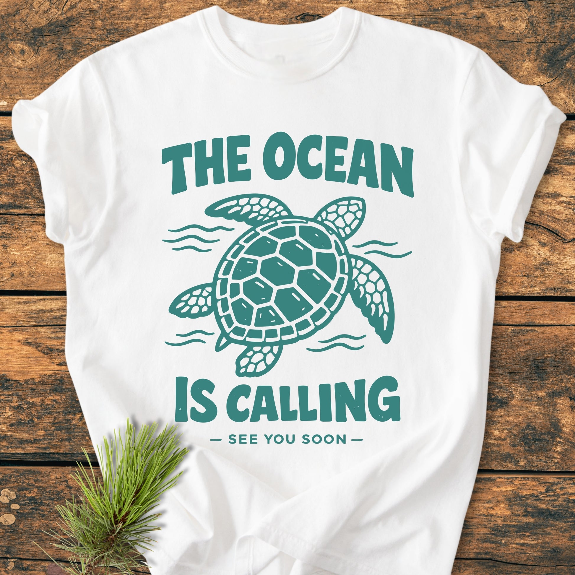 The Ocean Is Calling - Vintage Wash Tee features a teal sea turtle and waves graphic on a white T-shirt, laid on wood, with a pine sprig accentuating its relaxed, cozy design.