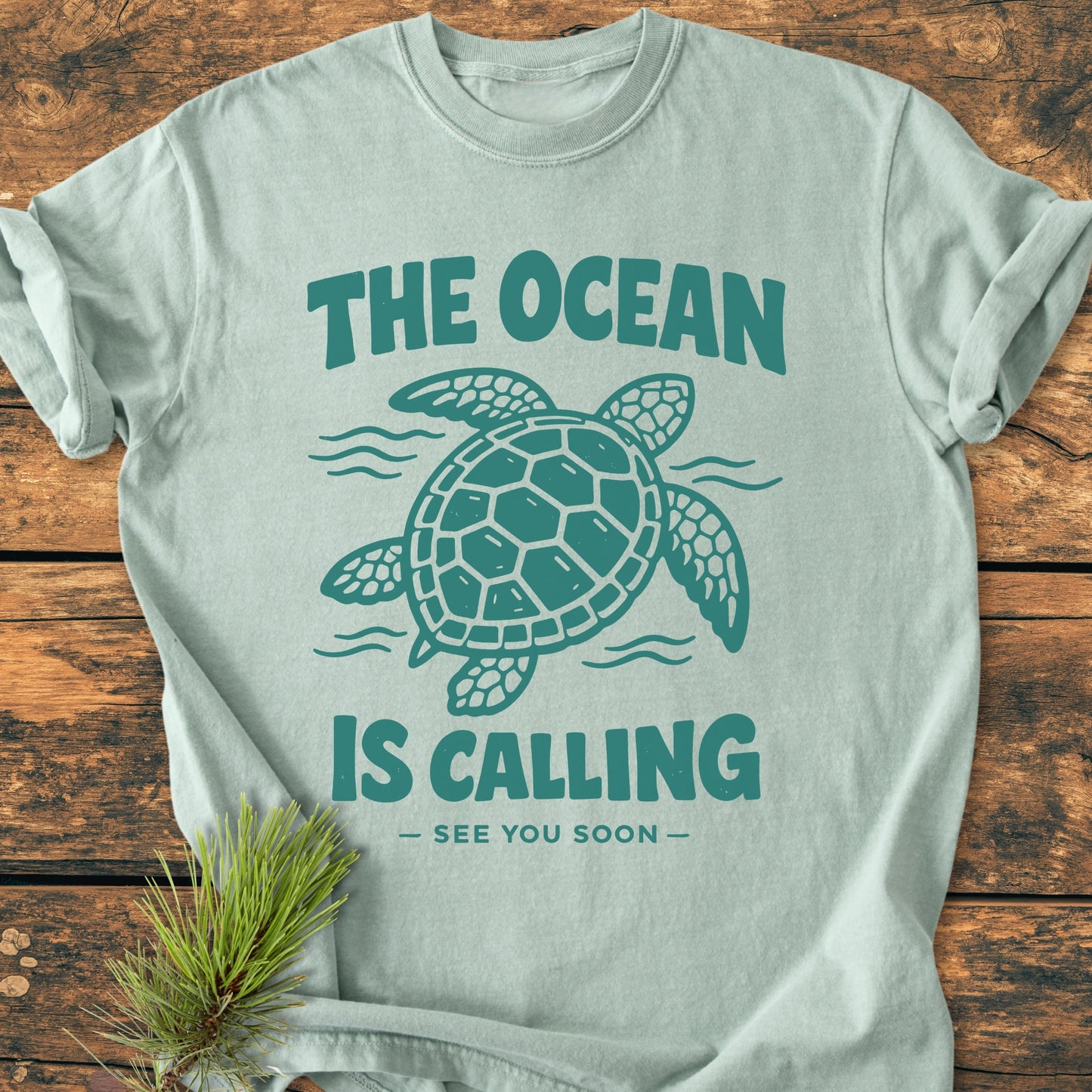 The Ocean Is Calling - Vintage Wash Tee features a sea turtle design with the slogan, displayed on a wooden surface. A pine branch accents the soft, cozy, garment-dyed fabric.