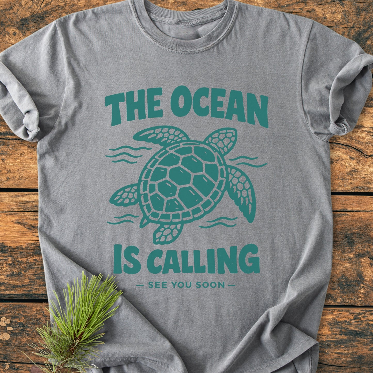 Heather-gray The Ocean Is Calling - Vintage Wash Tee featuring a sea turtle design and slogan, laid on rustic wood with a pine sprig; soft, durable, relaxed fit, 100% ring-spun cotton.