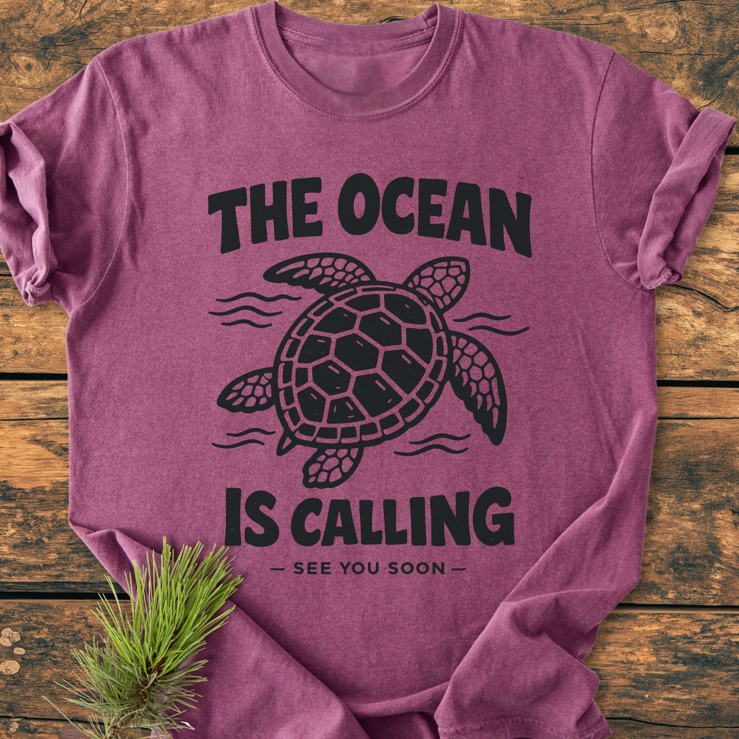 The Ocean Is Calling - Vintage Wash Tee features a stylized sea turtle design with wavy lines and slogan on a relaxed-fit, garment-dyed shirt, laid flat with a pine sprig accent.