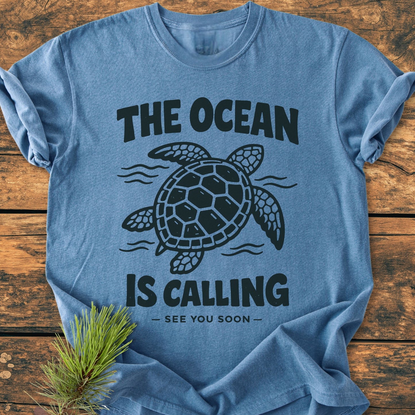 The Ocean Is Calling - Vintage Wash Tee displays a sea turtle and waves illustration on heather-blue fabric, laid on wood. Features include a rustic pine sprig accent and inspiring text.