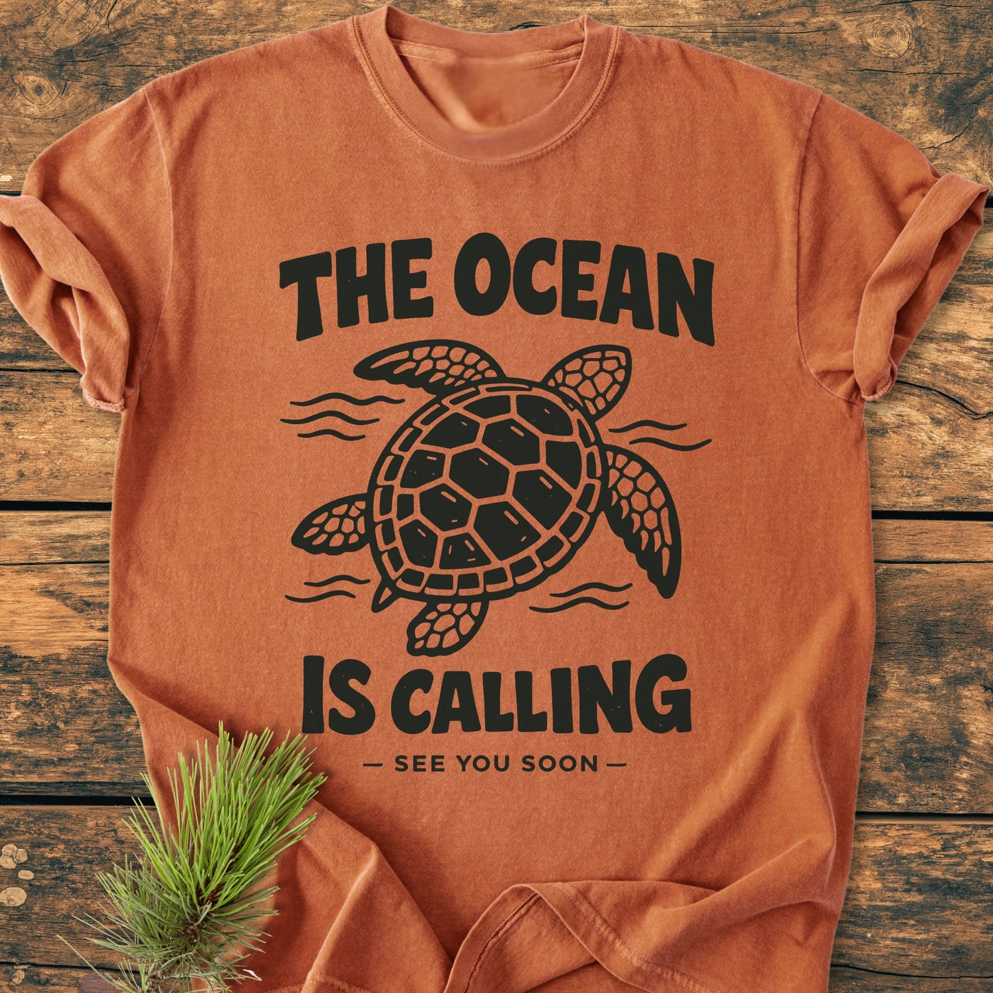 The Ocean Is Calling - Vintage Wash Tee features a sea turtle design with slogans, laid on wooden surface, paired with a pine sprig. Made from soft, relaxed-fit, garment-dyed cotton.