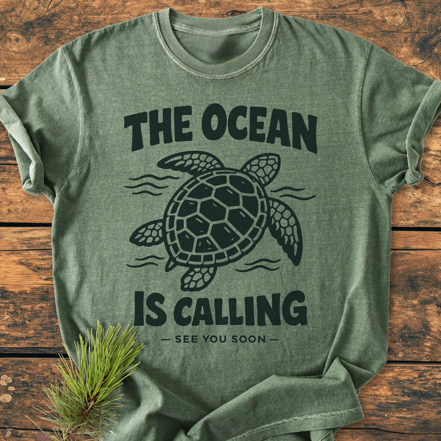 The Ocean Is Calling - Vintage Wash Tee features a sea turtle design with the slogan, laid on a wooden surface with pine, emphasizing its relaxed fit and soft Comfort Colors fabric.