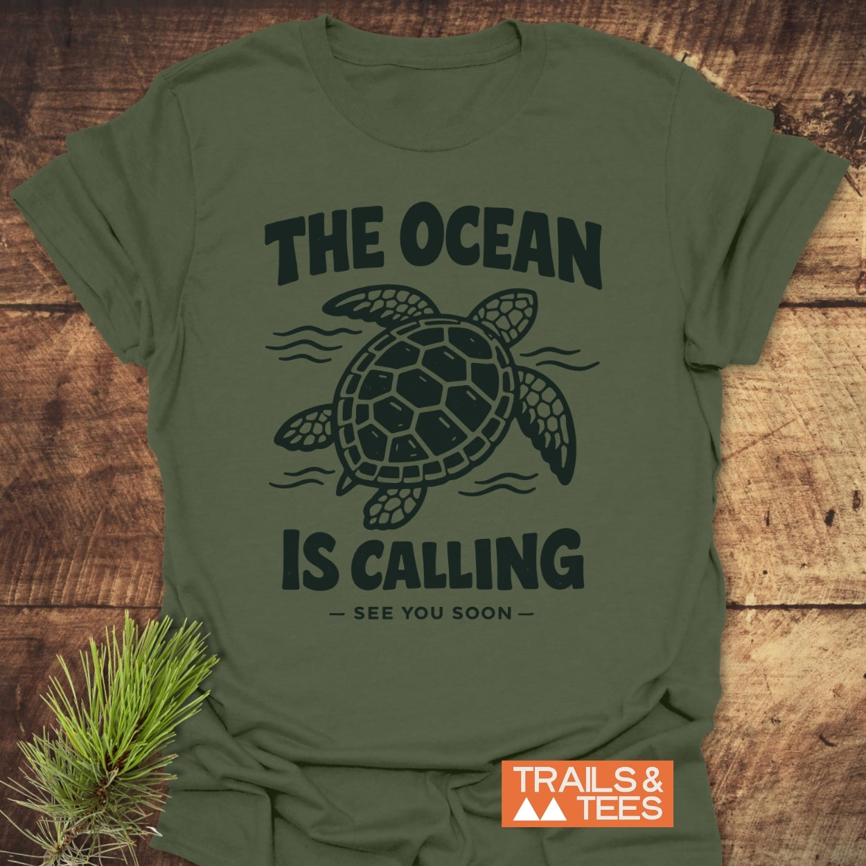 The Ocean Is Calling T-Shirt features a sea turtle illustration and slogan on olive-green fabric, styled with a pine sprig, showcasing its adventure-ready design by Trails & Tees.