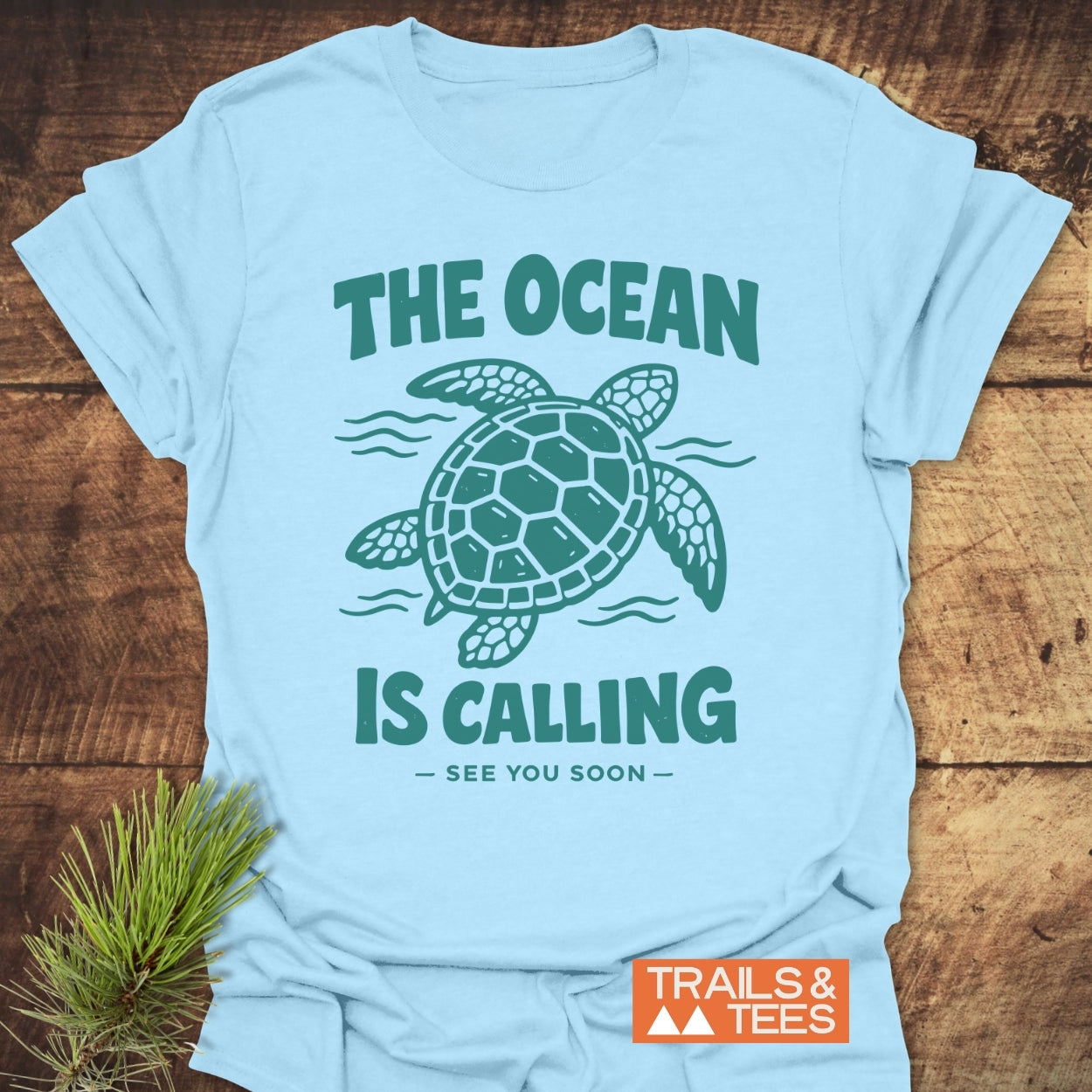 The Ocean Is Calling T-Shirt features a sea turtle and slogan, laid on wood with a pine sprig. Crafted from soft ring-spun cotton, it's perfect for outdoor enthusiasts.