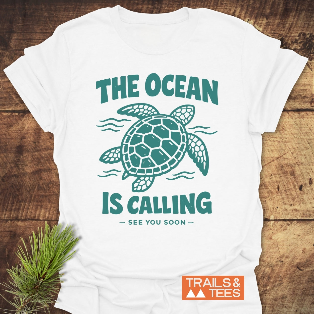 The Ocean Is Calling T-Shirt features a teal sea turtle design with waves and text. Laid on wood, it embodies outdoor spirit with soft, durable cotton, perfect for adventure enthusiasts.