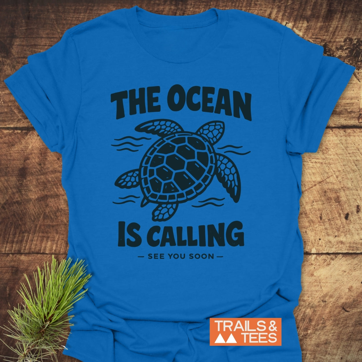 The Ocean Is Calling T-Shirt features a bold sea turtle design and slogan on soft, lightweight cotton, laid on a rustic surface, beside pine needles and a TRAILS & TEES logo.