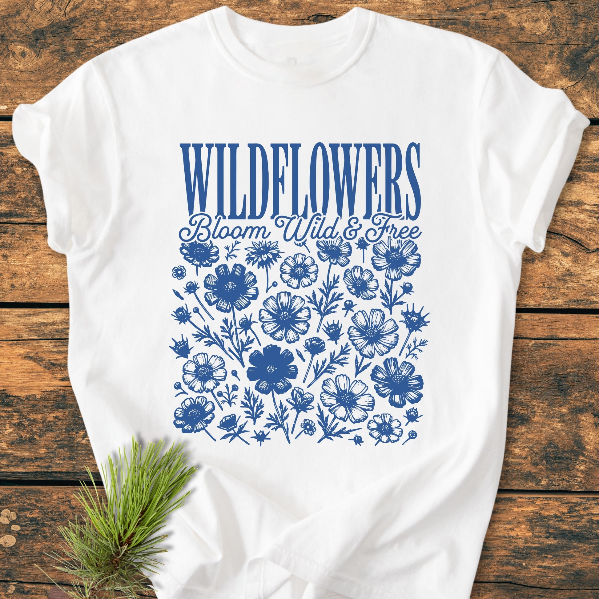 Wildflowers Bloom Wild And Free - Vintage Wash Tee features blue wildflower illustrations and text on a white T-shirt, laid flat on wood, with a pine sprig beside it for a rustic touch.