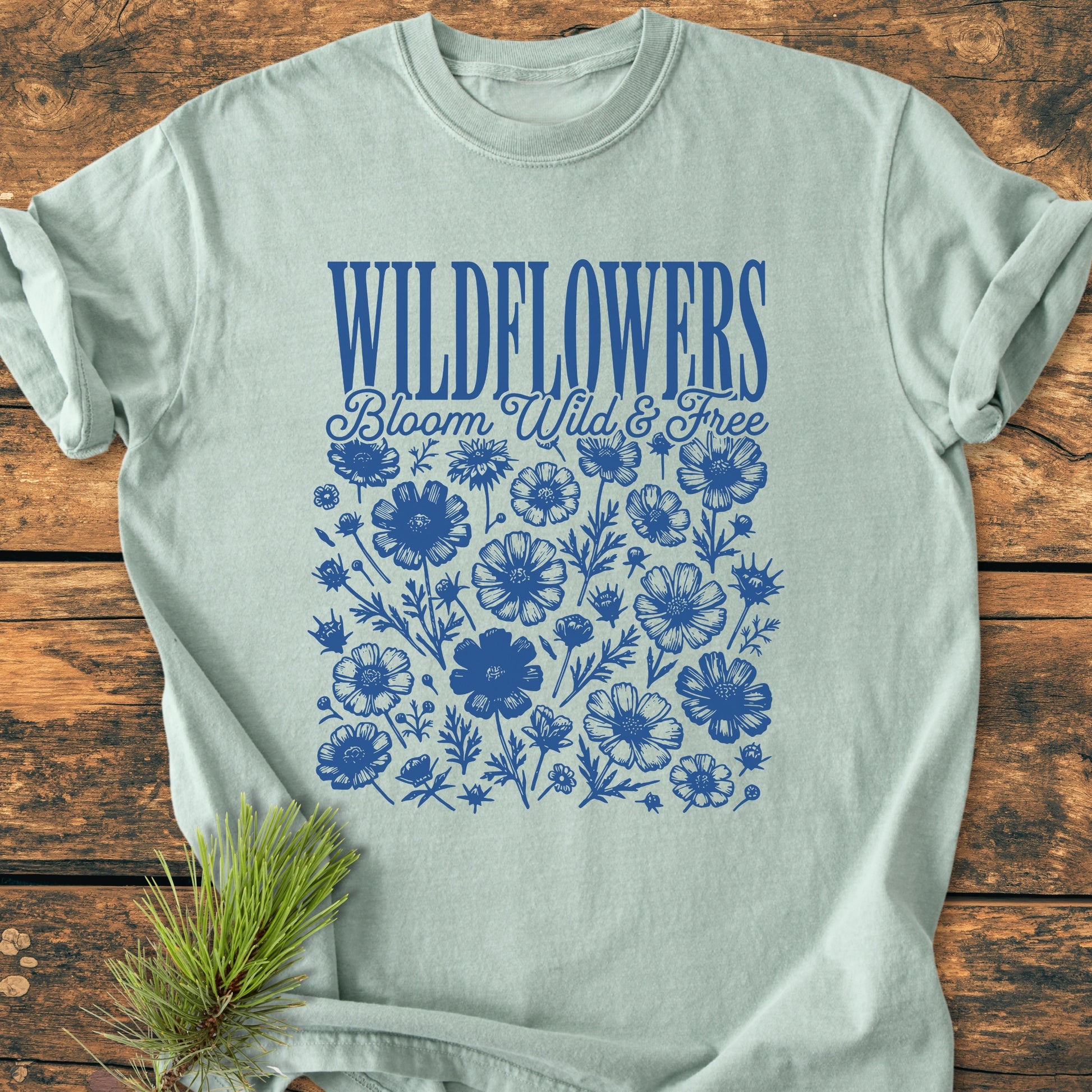 Wildflowers Bloom Wild And Free - Vintage Wash Tee features a blue WILDFLOWERS graphic with floral illustrations on a pale sage-green T-shirt, styled with a pine sprig.