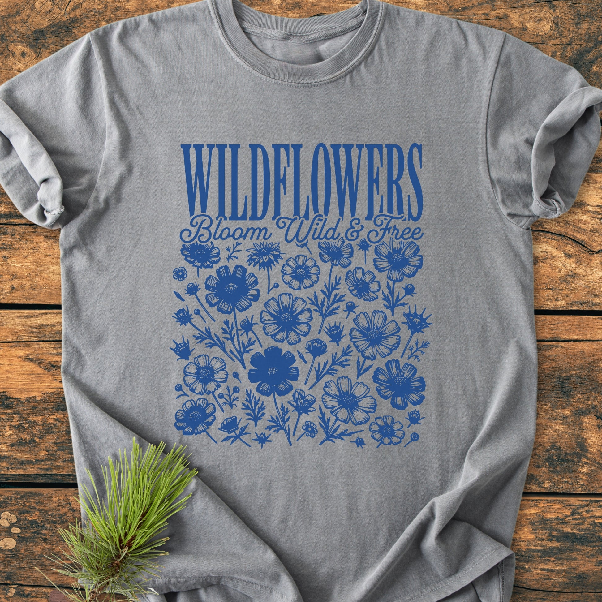Wildflowers Bloom Wild And Free - Vintage Wash Tee features royal blue WILDFLOWERS text and wildflower silhouettes on heather-gray fabric, laid flat on a wooden surface with a pine sprig accent.