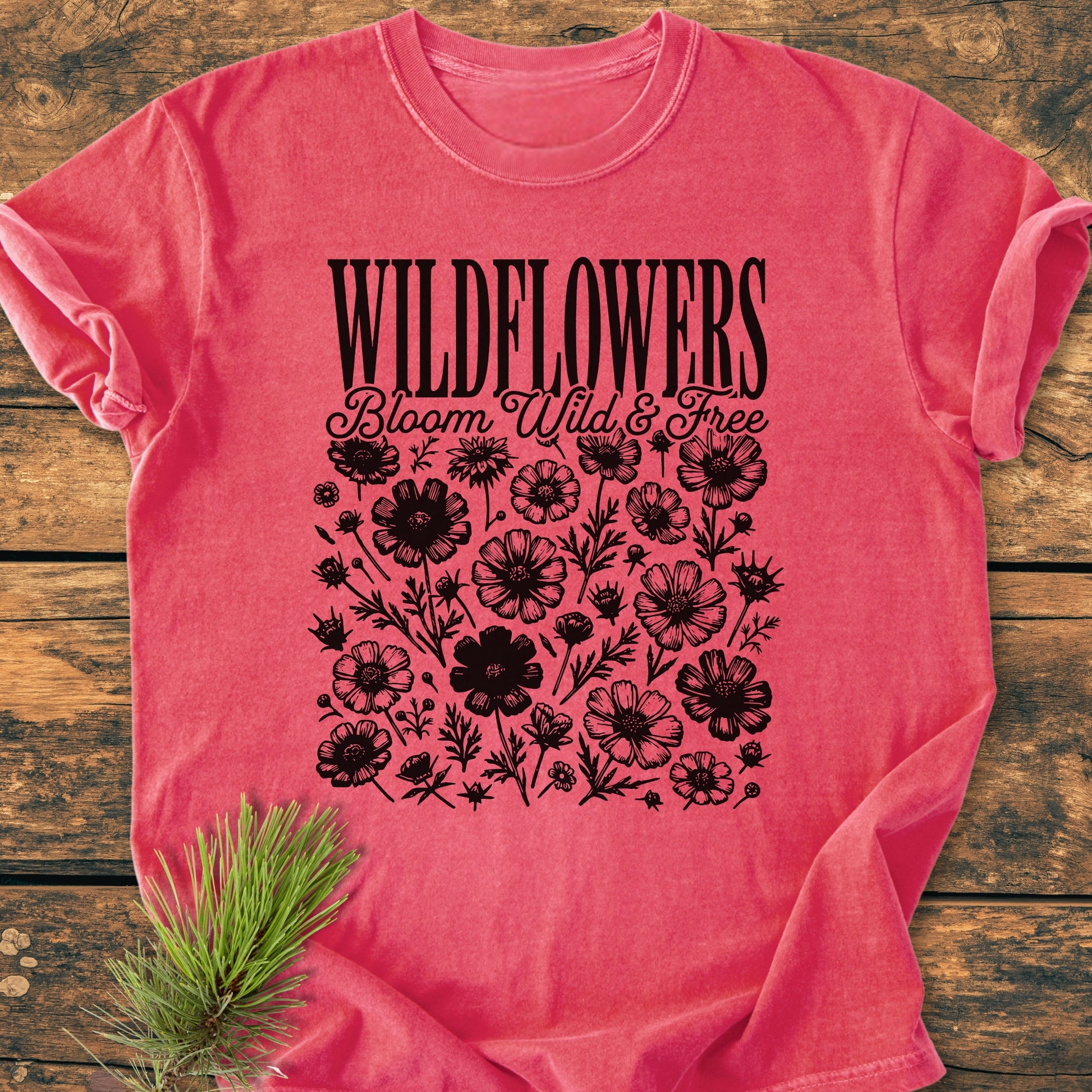 Wildflowers Bloom Wild And Free - Vintage Wash Tee on rustic wooden surface, featuring black wildflower graphics and text. Includes a small sprig of pine for a natural touch, emphasizing the tee's relaxed, cozy appeal.
