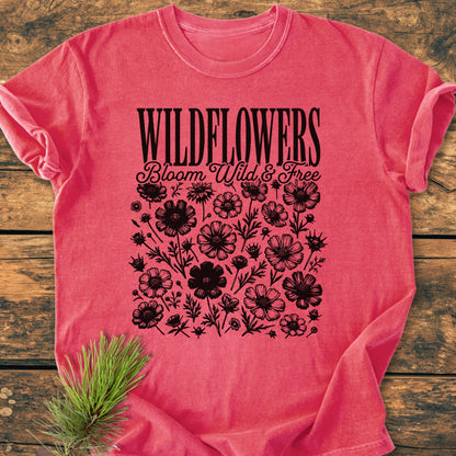 Wildflowers Bloom Wild And Free - Vintage Wash Tee on rustic wooden surface, featuring black wildflower graphics and text. Includes a small sprig of pine for a natural touch, emphasizing the tee's relaxed, cozy appeal.
