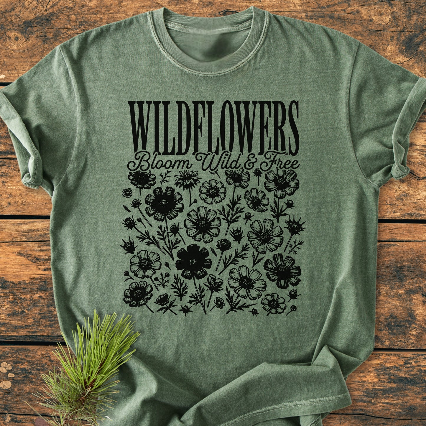Wildflowers Bloom Wild And Free - Vintage Wash Tee: Green crew-neck T-shirt with WILDFLOWERS and Bloom Wild & Free text above wildflower illustrations, styled with a pine sprig on a wooden surface.