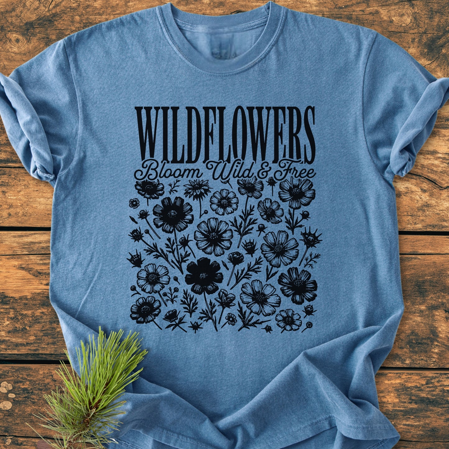 Wildflowers Bloom Wild And Free - Vintage Wash Tee, featuring black WILDFLOWERS text and wildflower illustrations on a heather-blue T-shirt, laid flat with a pine sprig beside it.