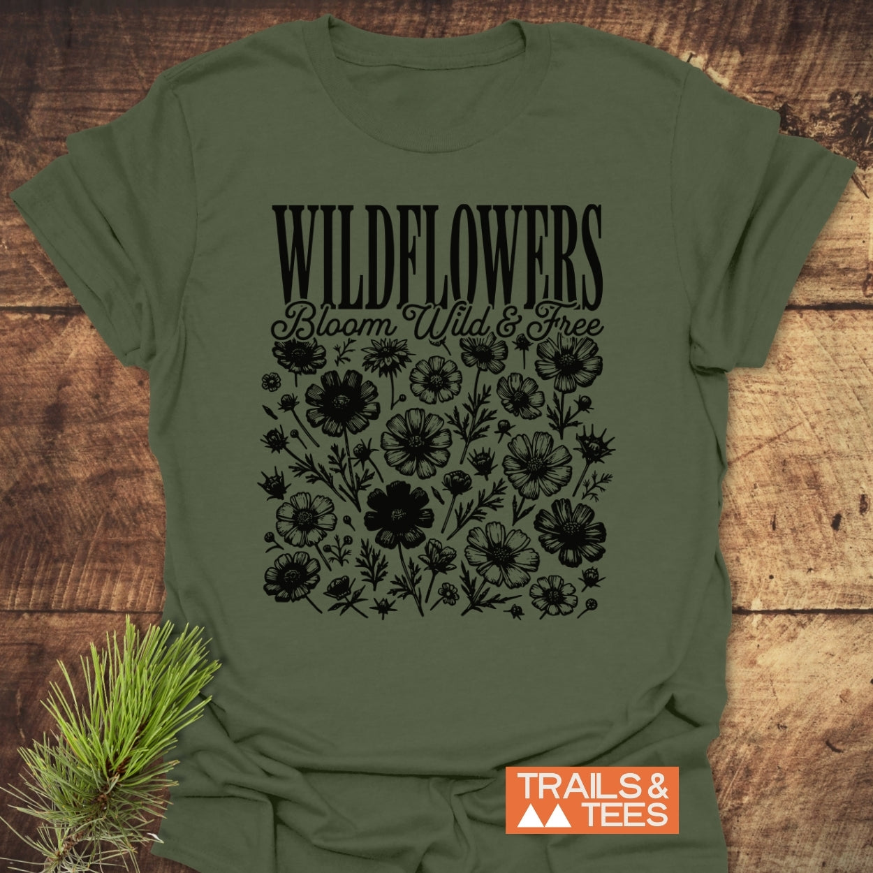 Wildflowers Bloom Wild And Free T-Shirt with “WILDFLOWERS” and wildflower silhouettes printed on olive-green fabric. Trails & Tees logo present, emphasizing an adventure-ready design.