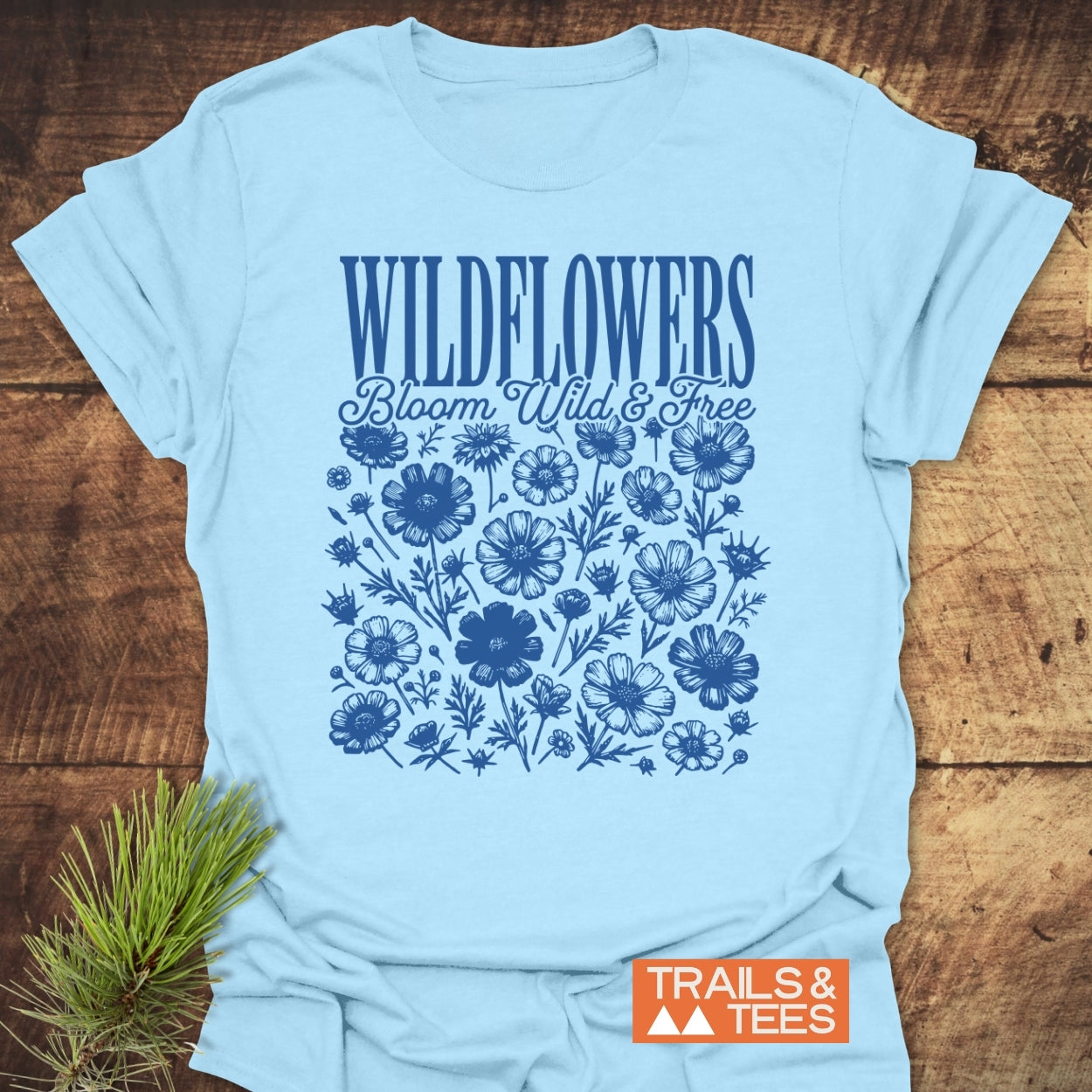 Wildflowers Bloom Wild And Free T-Shirt featuring dark blue WILDFLOWERS text, script Bloom Wild & Free, and wildflower illustrations. Includes TRAILS & TEES logo, styled with pine sprigs for an outdoor feel.