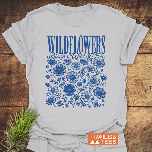 Wildflowers Bloom Wild And Free T-Shirt laid flat on rustic wood, featuring blue “WILDFLOWERS” text, script “Bloom Wild & Free,” and hand-drawn wildflowers; pine sprig nearby, “TRAILS & TEES” logo in corner.