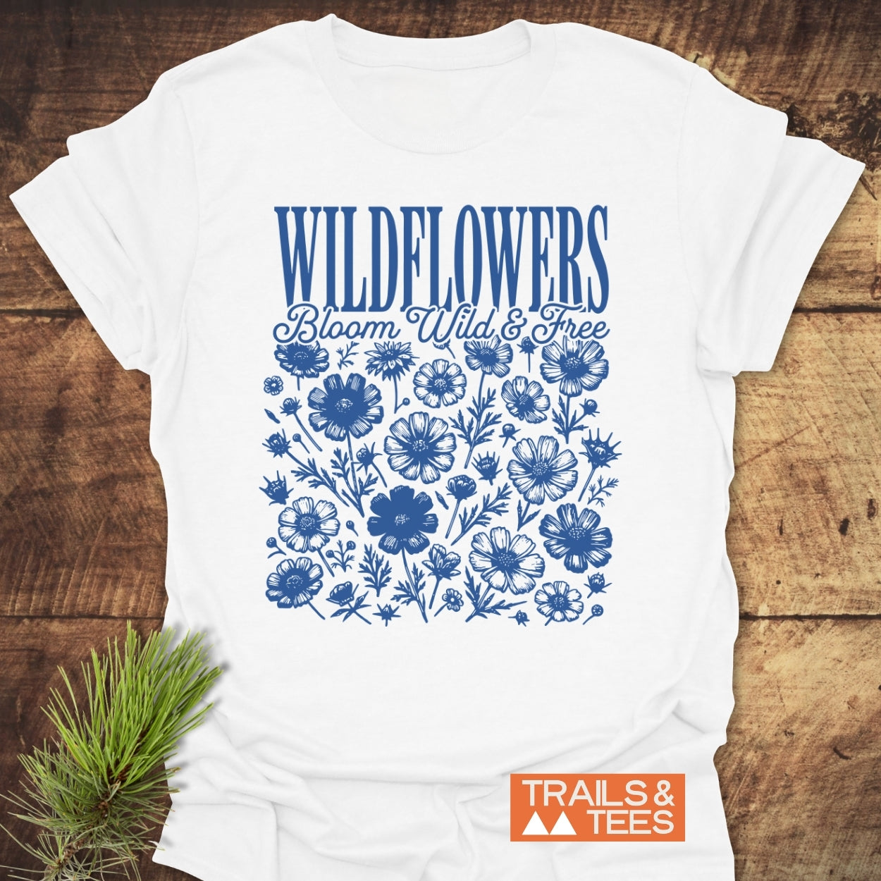 Wildflowers Bloom Wild And Free T-Shirt featuring a blue wildflower graphic and inspirational text on a white tee, laid on wood with a pine branch beside it.