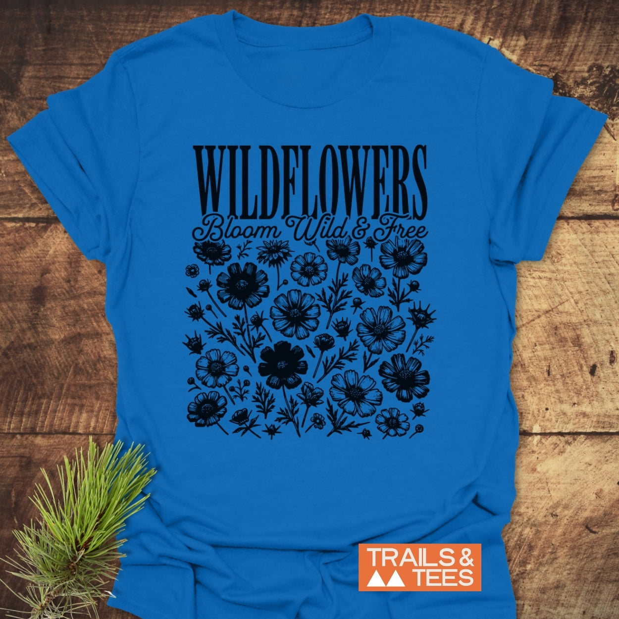 Wildflowers Bloom Wild And Free T-Shirt featuring hand-drawn wildflowers and text in black on a cobalt-blue fabric, styled with a pine sprig, showcasing trail-tested comfort and outdoor spirit.