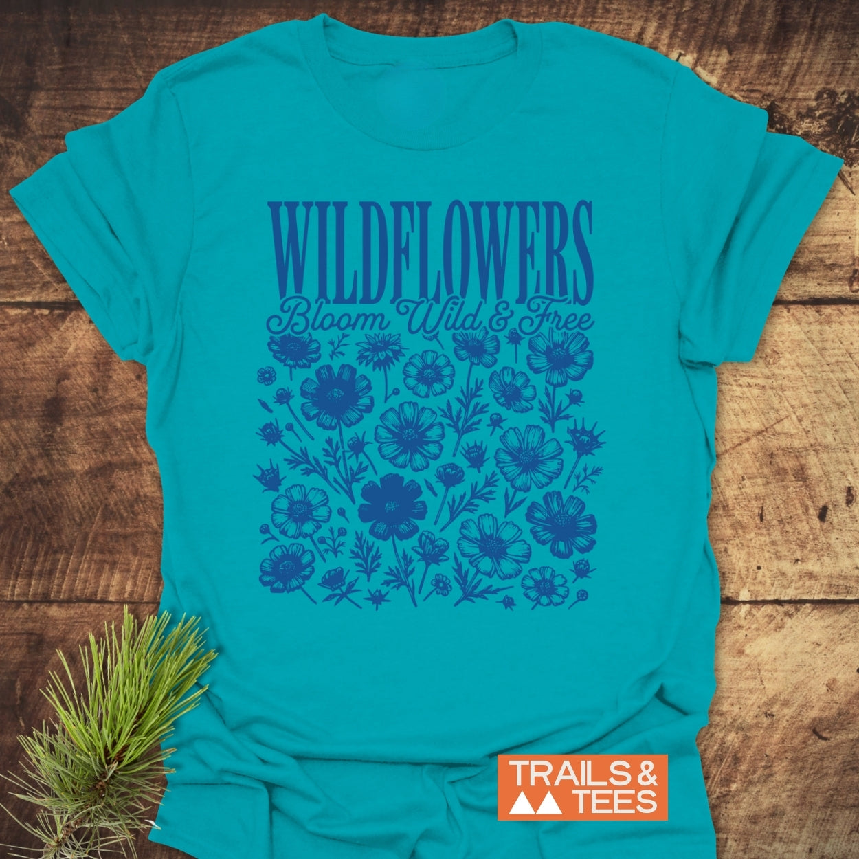 Teal Wildflowers Bloom Wild And Free T-Shirt with dark blue wildflower graphic, “TRAILS & TEES” logo, and a pine branch accent, showcasing comfort and outdoor-inspired design.