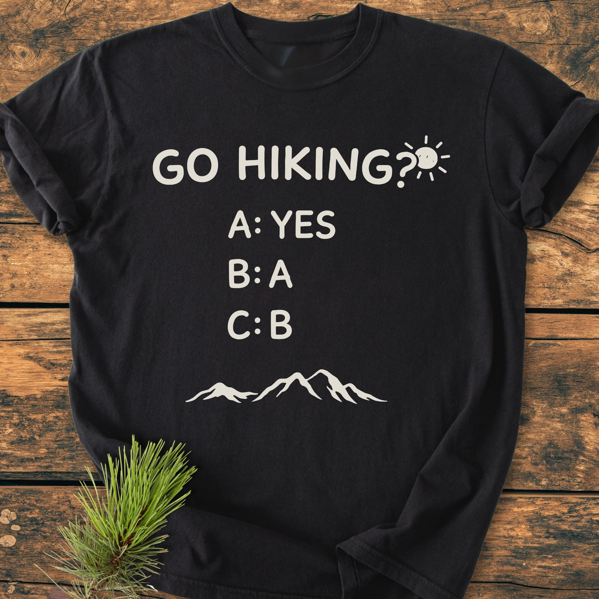 Go Hiking? - Vintage Wash Tee features a playful slogan with sun and mountain graphics on a black, soft-washed T-shirt, laid on a wooden surface with a pine sprig.