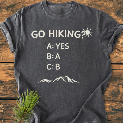 Go Hiking? - Vintage Wash Tee displayed flat on wood, featuring off-white slogan and mountain graphic. Rustic charm enhanced by a pine sprig, showcasing relaxed fit and garment-dyed texture.