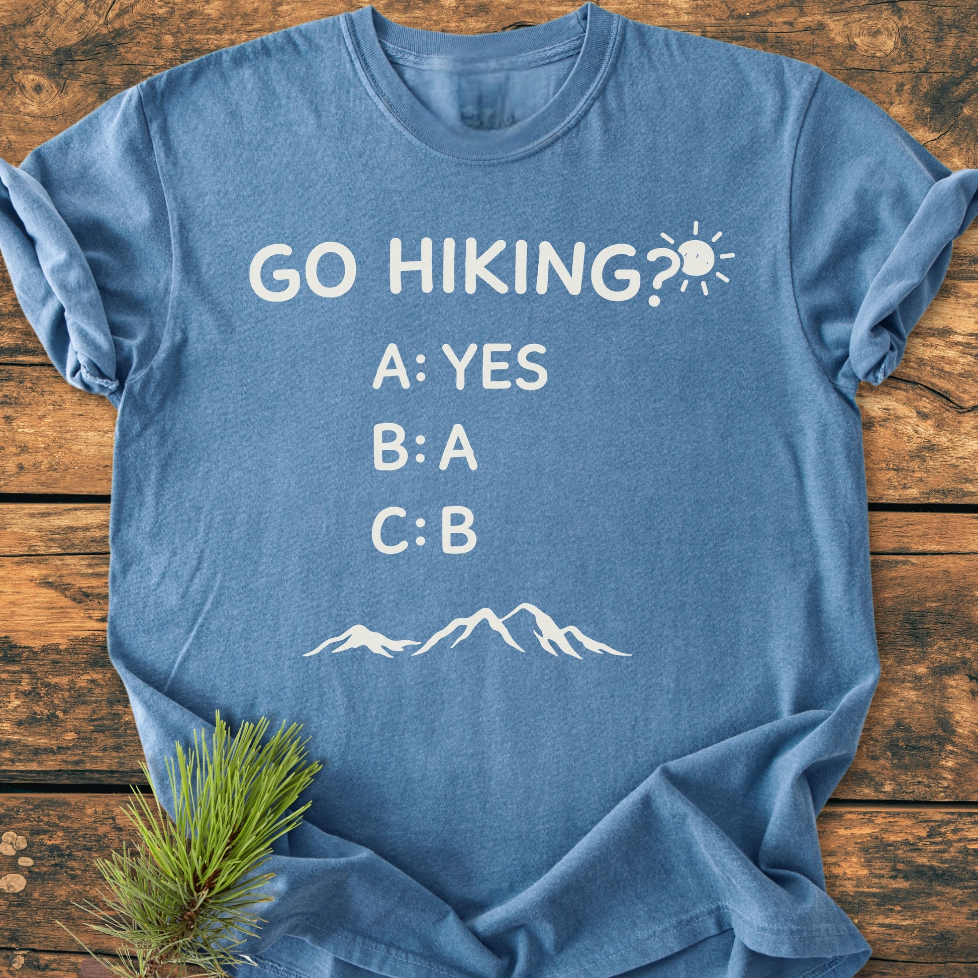 Go Hiking? - Vintage Wash Tee flat-lay on rustic wood, featuring playful hiking text and mountain outline, emphasizes comfort and durability with its relaxed fit and garment-dyed, ring-spun cotton fabric.