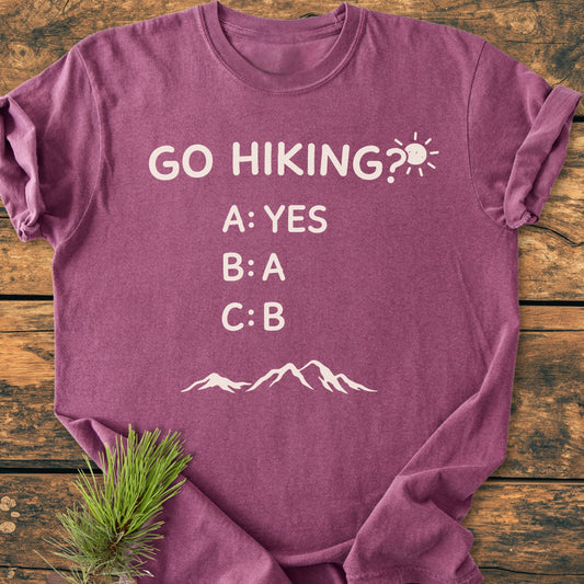 Heathered plum Go Hiking? Vintage Wash Tee with playful hiking-themed graphic and mountain silhouette, displayed flat-lay style on wood. Includes pine needles, showcasing its cozy, durable, and relaxed fit design.