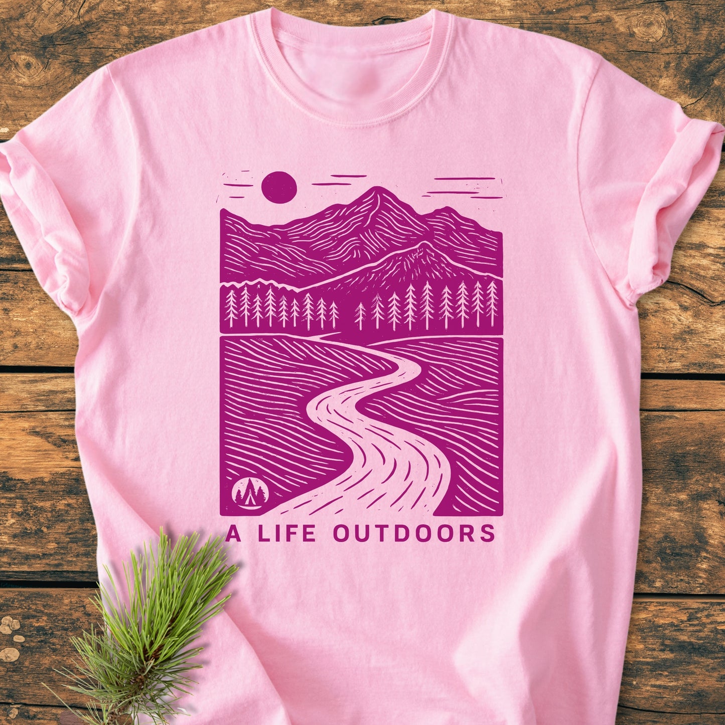 A Life Outdoors - Vintage Wash Tee displays a scenic, magenta outdoor print on soft-pink fabric, laid on wood with a pine branch. Relaxed fit, durable stitching, perfect for daily wear.