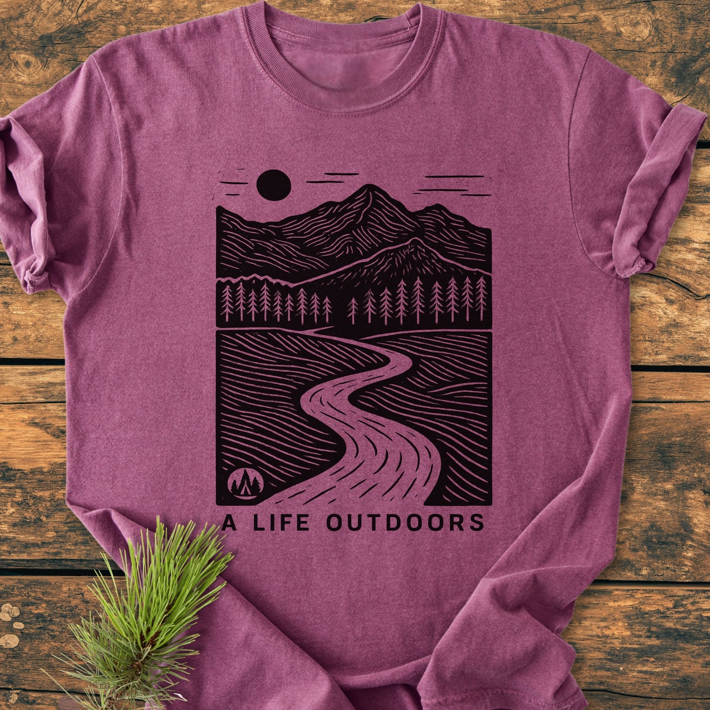 A Life Outdoors - Vintage Wash Tee features a mountain landscape illustration with sun, pine trees, and river on a heathered mauve T-shirt, complemented by a rustic wooden backdrop and pine branch accent.