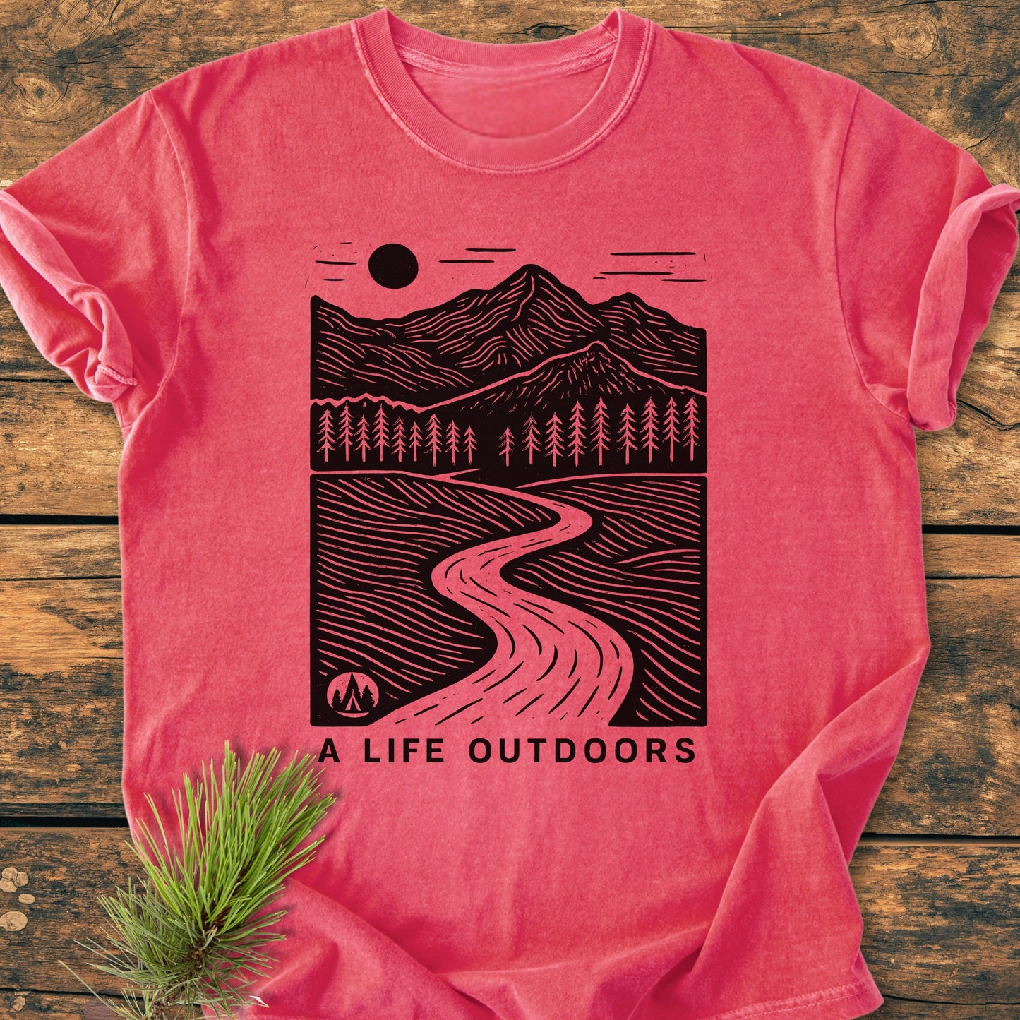 A Life Outdoors - Vintage Wash Tee features a printed linocut-style landscape with mountains and pines on a heathered red-pink background, styled with a pine sprig on a wooden surface.