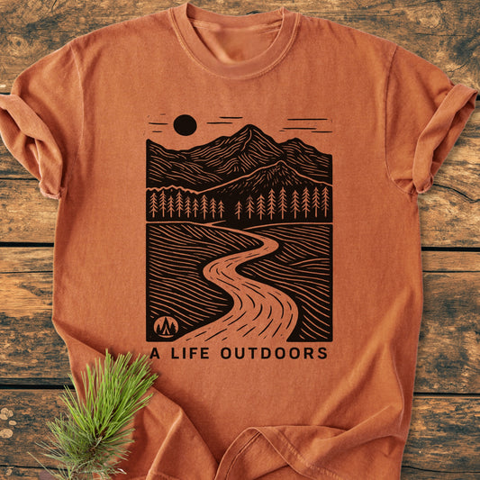 A Life Outdoors - Vintage Wash Tee: Burnt-orange T-shirt with a black wood-cut-style illustration of mountains, trees, and path, slogan beneath. Laid on wood, pine sprig beside.
