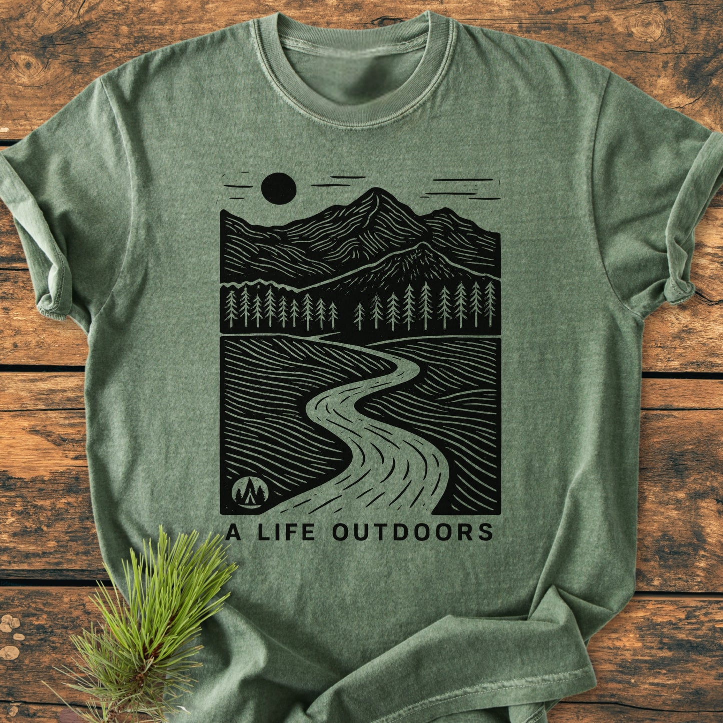 A Life Outdoors - Vintage Wash Tee features a hand-drawn landscape with river, trees, and mountains on heather-green fabric, laid on wood with a pine twig. Cozy, relaxed fit.