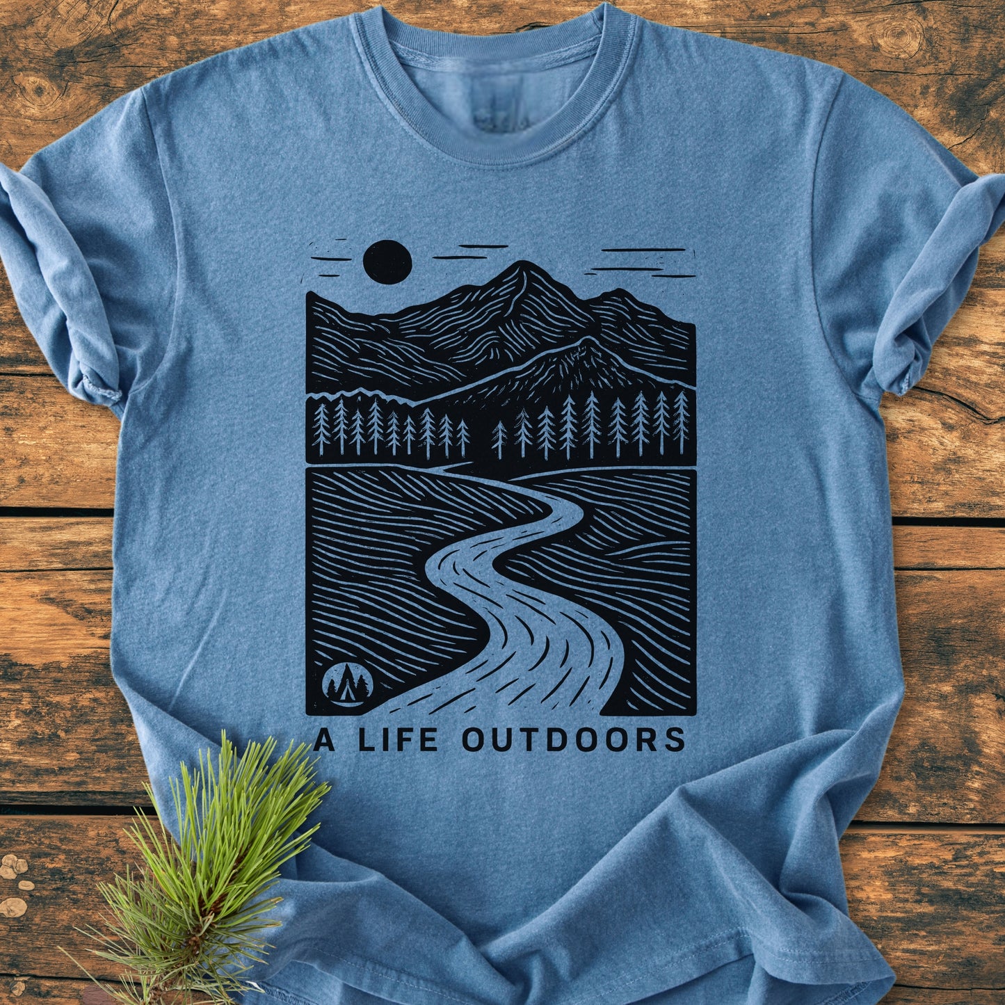A Life Outdoors - Vintage Wash Tee features a mountain graphic with trees, path, sun, and tent, styled with a pine sprig, emphasizing outdoor adventure on a heather-blue T-shirt.