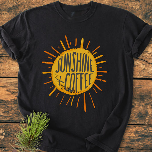 Sunshine + Coffee - Vintage Wash Tee featuring a sunburst design on a black shirt, laid on wood with a pine sprig. Made from soft, durable, pre-shrunk, ring-spun cotton.