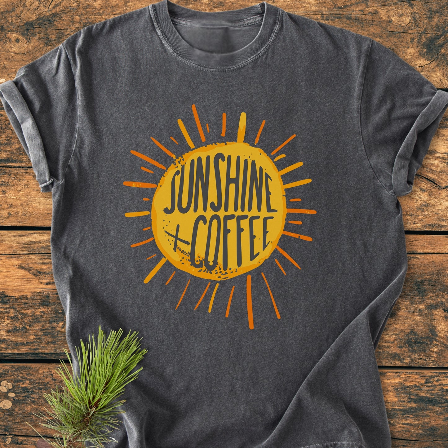 Sunshine + Coffee - Vintage Wash Tee features a dark-gray design with a vibrant sunburst graphic and text, laid flat on a wooden surface with a pine needle sprig accent.