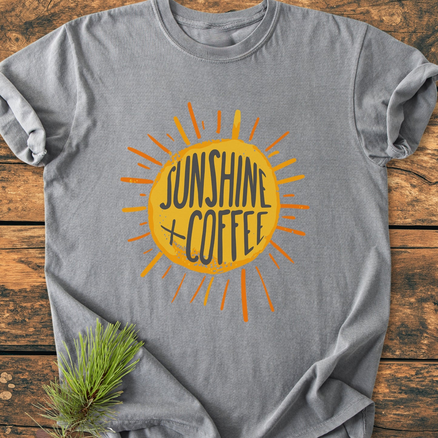 Sunshine + Coffee - Vintage Wash Tee: Heather-gray T-shirt with a sun graphic and text, laid on wood. Features relaxed fit, durable stitching, and soft-washed cotton for comfort. Pine sprig beside it.