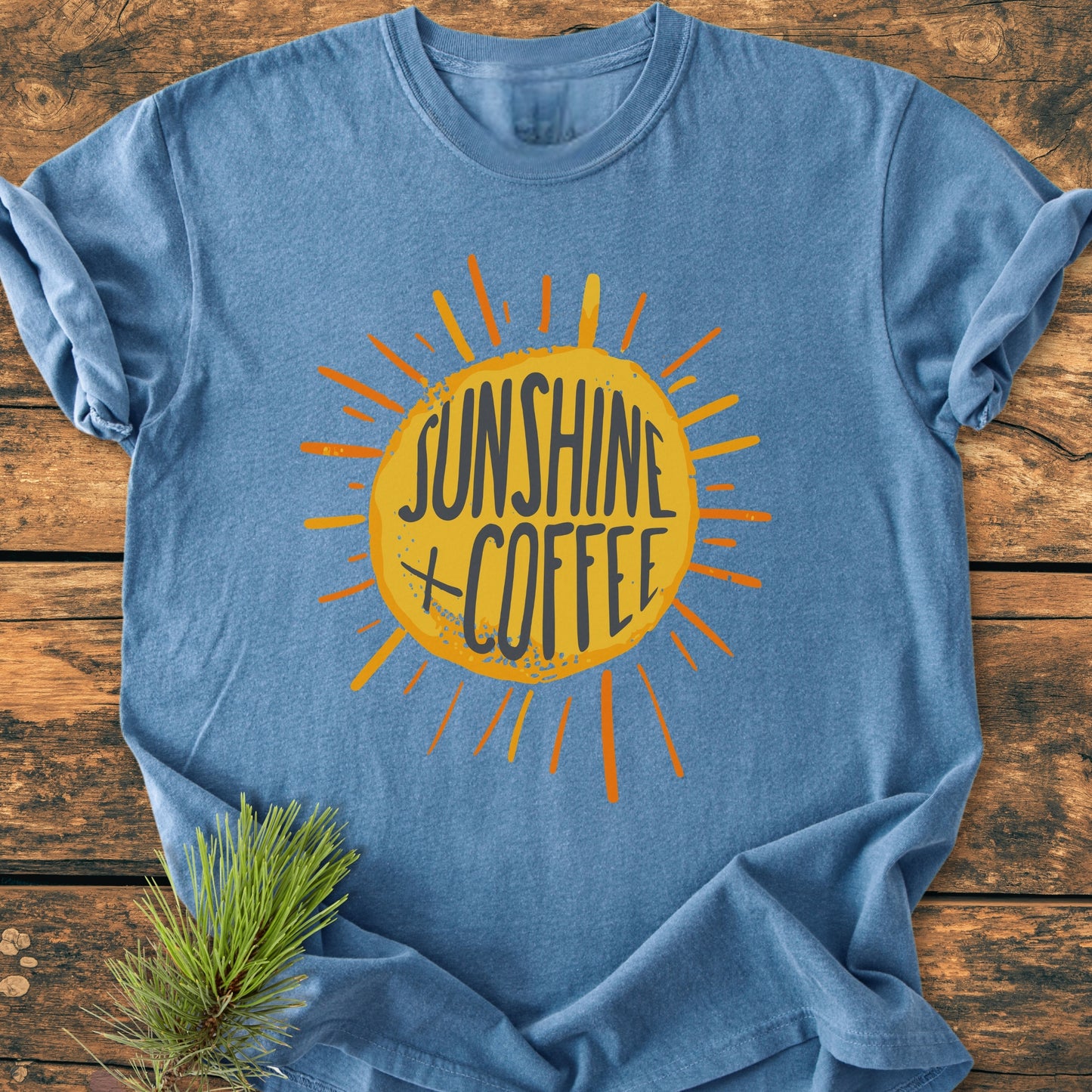 Sunshine + Coffee - Vintage Wash Tee laid on rustic wood, featuring a yellow sunburst with orange rays. A sprig of evergreen accents the scene, highlighting the tee's relaxed, cozy style.
