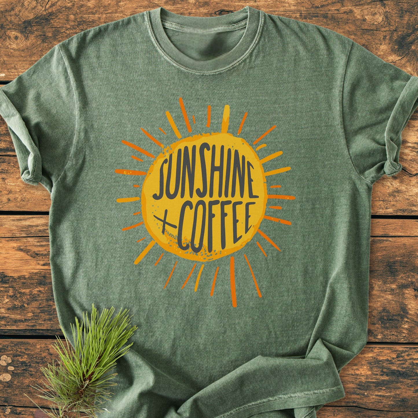 Sunshine + Coffee - Vintage Wash Tee in heather-green, featuring a yellow sunburst graphic and text, laid flat on wood. Pine sprig nearby. Relaxed fit, soft-washed cotton for comfort.