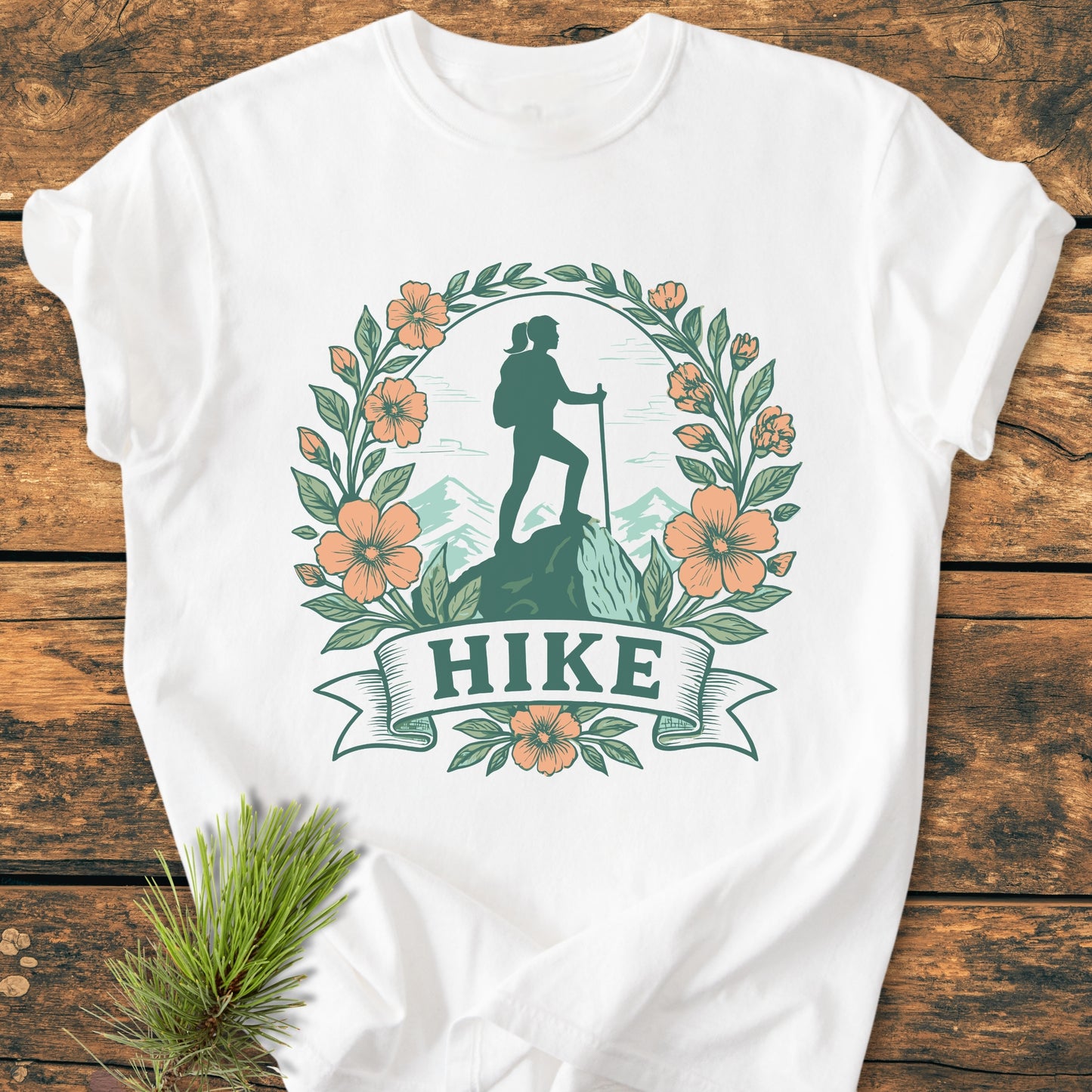 HIKE Flower - Vintage Wash Tee features a stylized hiker design with mountain backdrop, framed by leaves and flowers, on a white T-shirt laid on wood, accompanied by a pine sprig.