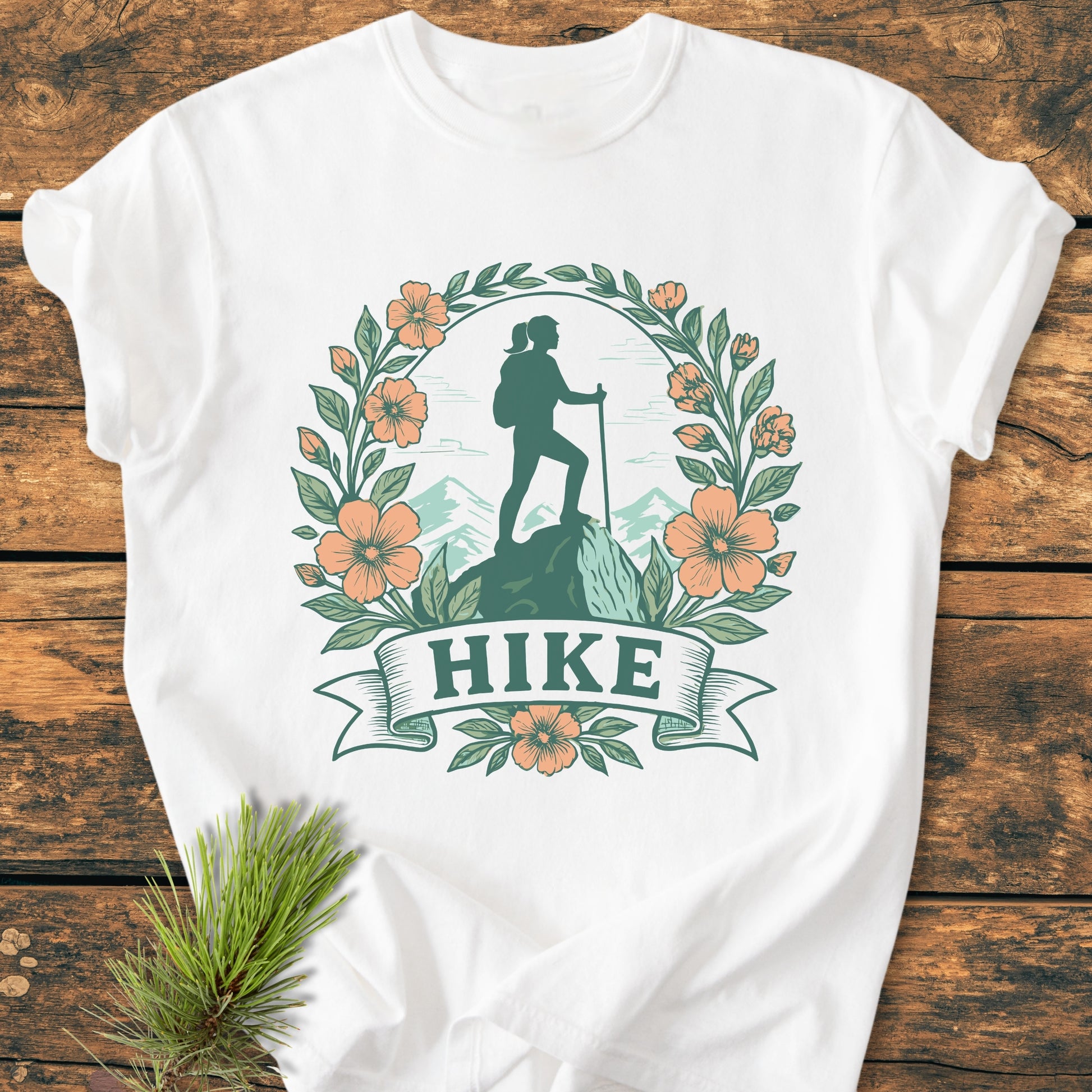 HIKE Flower - Vintage Wash Tee features a stylized hiker design with mountain backdrop, framed by leaves and flowers, on a white T-shirt laid on wood, accompanied by a pine sprig.